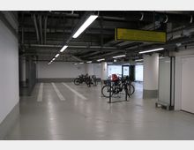 Unterirdischer Fahrradabstellraum mit mehreren angelehnten Fahrrädern, beleuchtet durch helle Deckenlampen. Die Umgebung zeigt graue Wände, technische Rohrleitungen an der Decke und eine Ausfahrt mit Glastüren im Hintergrund.