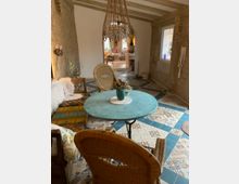 Gemütlicher Innenraum mit rustikalem Charme, bestehend aus einer Sitzecke mit einem runden, türkisfarbenen Tisch, geflochtenen Stühlen und gemusterten Fliesenboden. Die Wände sind aus grobem Putz und Stein, ein großer Kronleuchter hängt von der Balkendecke, und im Hintergrund ist ein weiterer Wohnbereich sichtbar.