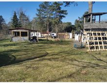 Ein ländlicher Garten mit Rasenfläche, einer kleinen Holzhütte links im Bild sowie weiteren Gebäuden und Wohnwägen im Hintergrund. Rechts steht ein hölzerner Unterstand mit Kletterelementen, umgeben von hohen Bäumen und einer friedlichen Waldkulisse.