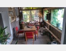 Veranda mit rustikalem Interieur, einem Holztisch und roten Stühlen in der Mitte. Fensterfronten mit Blick ins Grüne, Topfpflanzen und diverse Gegenstände wie ein Fahrrad und Holzkisten dekorieren den Raum.