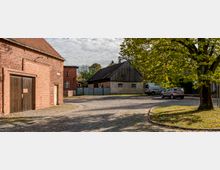 Gepflasterter Hof mit einem roten Backsteingebäude links und einem einfachen Gebäude mit verputzten Wänden und Holzdach in der Mitte. Im Vordergrund steht ein großer Baum mit herbstlichen Blättern, daneben parken zwei Autos auf dem Platz.