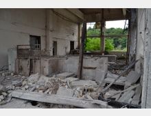 Ruine eines verlassenen Industriegebäudes mit zerfallenem Beton, freiliegenden Stahlstrukturen und Trümmern am Boden. Große Fensteröffnungen geben den Blick auf üppige, grüne Vegetation außerhalb frei.