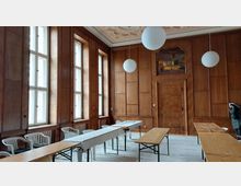 Raum mit holzvertäfelten Wänden, mehreren hohen Fenstern auf der linken Seite und einer verzierten Decke mit kugelförmigen Hängelampen. In der Mitte stehen einfache Holztische und Kunststoffstühle, an der Stirnwand befindet sich eine Holztür mit einem Gemälde darüber.