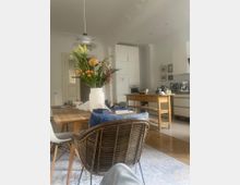 Heller Wohn- und Essbereich in einer Wohnung mit hohen Decken und Stuckverzierungen. Im Vordergrund steht ein Esstisch aus Holz mit einem Blumenstrauß, im Hintergrund sind eine moderne Küche mit weißen Schränken und ein Holzregal zu sehen.