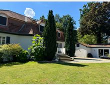 Weißes Einfamilienhaus mit roten Dachziegeln und mehreren Dachgauben, umgeben von einem gepflegten Garten mit Rasenfläche, Sträuchern und hohen Bäumen. Eine halbrunde Steintreppe führt zum Hauseingang, daneben ein kleiner gepflasterter Hof.