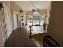 Halbkreisförmiger Raum mit großen Bogenfenstern, die auf eine Terrasse mit weißem Geländer blicken, hellem Holzfußboden, eleganter Treppe mit geschwungenem Metallgeländer und weißen Türen mit dekorativen Zierleisten.