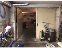 Ein Kellerraum mit sichtbaren Ziegelwänden und niedrigem Betondecken. Im Vordergrund befinden sich ein Fahrrad, ein Staubsauger und diverse Gegenstände, im Hintergrund sind Leitern und Lagerkisten erkennbar.