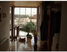 Ein Flur mit Fensterfront, durch die eine verschneite Landschaft sichtbar ist. In der Mitte stehen mehrere Topfpflanzen auf glänzendem, gefliestem Boden, rechts sind eine Garderobe mit Jacken und links eine Heizung zu sehen.