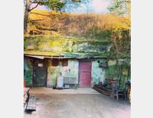 Ein rustikaler Hofbereich vor einer grob verputzten Wand mit verwitterten Türen, eine in Rot und eine in Dunkelblau. Im Vordergrund stehen eine Holzbank, ein Metallbehälter und verschiedene Werkzeuge; im Hintergrund ist eine mit Moos bewachsene Erdwand und Bäume erkennbar.