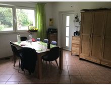 Esszimmer mit großem rechteckigem Holztisch, umgeben von mehreren Stühlen. Im Raum befinden sich ein Kleiderschrank und Kommode ebenfalls aus Holz, ein großes Fenster mit grünem Vorhang sowie eine Tür mit Glaseinsätzen.