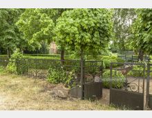 Ein gepflegter Garten mit hohen Bäumen, geschnittenen Hecken und schmiedeeisernem Zaun samt offenem Tor. Im Hintergrund ist eine Holzgitterbank und ein altes Ziegelgebäude zu erkennen.