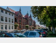 Straßenszene mit einem historischen roten Backsteingebäude mit verziertem Giebel und kleinen Türmchen, flankiert von einem weißen Gebäude mit Blumenkästen an den Fenstern. Im Vordergrund geparkte Autos und auf der rechten Seite Bäume, die Schatten spenden.