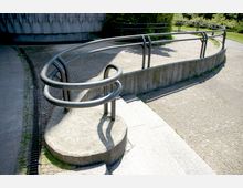 Eine gebogene Rampe mit Edelstahlgeländer führt von einem gepflasterten Weg hinauf zu einer erhöhten Fläche aus Beton. Im Hintergrund sind Vegetation und ein Gebäude mit grauer Betonfassade sichtbar.