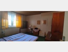 Schlicht eingerichtetes Schlafzimmer mit einem Bett im Vordergrund, einem Fenster mit gelben Vorhängen links und einem kleinen Tisch mit Lampe an der Wand. Die Wände sind hell, und Holzmöbel sowie Bilderrahmen dekorieren den Raum.