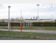 Pferderennbahn mit Tribünen im Hintergrund, flankiert von Flutlichtmasten und umgeben von Hecken und Wiesen. Im Vordergrund befindet sich eine Schranke mit einem Gehweg und einer Parkfläche.