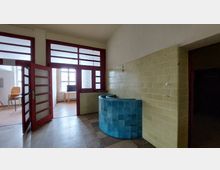 Ein Innenraum mit gelb gefliesten Wänden und einem hellblauen, halbrunden Fliesentresen. Im Hintergrund sind rote Türrahmen mit Glaselementen sowie ein Fenster mit Tageslicht zu sehen.