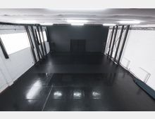 Ein leerer Sportraum mit schwarzem, glänzendem Boden, weißen Ziegelwänden und freiliegenden Holzbalken. Der Raum hat große Fenster an den Seiten und eine dunkle Wand mit einem schwarzen Vorhang im Hintergrund.