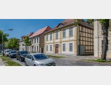 Straßenszene mit historischen Wohnhäusern, darunter ein gelbes Fachwerkhaus mit weißen Fenstern und roten Ziegeldächern. Geparkte Autos stehen entlang der gepflasterten Straße, und im Hintergrund sind Bäume sowie moderner Gebäudebestand sichtbar.