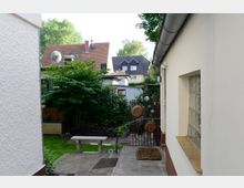 Schmaler Hinterhof mit gepflastertem Weg, links und rechts von Gebäuden begrenzt. Im Hintergrund ein kleiner Garten mit einer Sitzbank, Bäumen und Sträuchern, sowie angrenzenden Wohnhäusern mit Satteldächern.