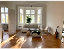 Helles Wohnzimmer mit hohen Decken, großen Sprossenfenstern und Parkettboden. Der Raum ist eingerichtet mit einem beigefarbenen Sofa, einem Sessel, einem Teppich, einem Spiegel, einer Gitarre an der Wand und einer Zimmerpflanze vor den Fenstern.