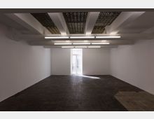 Leerer Innenraum mit weißen Wänden, einer hohen Glastür hinten in der Mitte und einem dunklen Boden aus kleinen Fliesen. Die Decke ist mit Beleuchtungsröhren und sichtbaren strukturellen Elementen gestaltet.