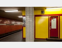 U-Bahn-Station mit gelben Wänden, roten Türrahmen und einer Säule mit weißen Kacheln. Auf der linken Seite ist eine rote Ziegelmauer mit dem Stationsnamen "Yorckstraße".