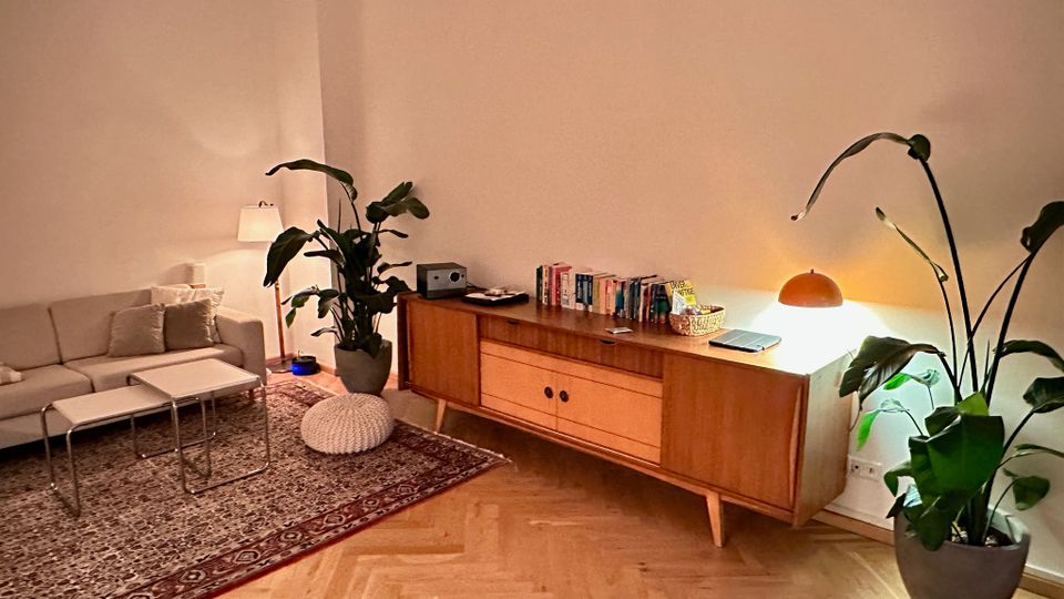 Gemütlicher Wohnbereich mit hellgrauem Sofa und zwei Couchtischen auf einem gemusterten Teppich sowie Parkett im Fischgrätmuster. Daneben steht ein niedriges Holz-Sideboard mit Büchern und Lampe, flankiert von großen Zimmerpflanzen vor einer schlichten weißen Wand.
