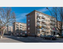 Wohngebäude an einer Straßenecke mit schlichtem Putz und Balkonen. Entlang der Straße parken Autos, und im Vordergrund stehen kahle Bäume; der Himmel ist klar und blau.