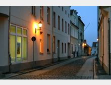 Schmale, kopfsteingepflasterte Gasse mit mehrstöckigen Wohnhäusern auf beiden Seiten bei Dämmerung. Warme Straßenlaternen beleuchten die Gebäude, deren Fenster teilweise Licht ausstrahlen.