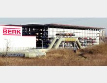 Ein modernes Industriegebäude mit der Aufschrift "BERK" neben einem offenen mehrstöckigen Parkhaus, in dem Autos auf mehreren Ebenen geparkt sind. Im Vordergrund befinden sich Rohre mit Graffiti, umgeben von trockenem Gras und Büschen.