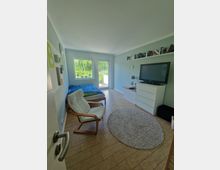 Helles Schlafzimmer mit Holzboden, einem Bett, einem weißen Sessel, einer Kommode und einem Fernseher. Große Fenster und eine Glastür bieten Blick auf grüne Vegetation; an der Wand befinden sich ein Regal mit Büchern und kleine Dekorationen.