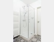 Eine moderne Duschkabine mit Glaswänden und einem Duschkopf mit verstellbarem Schlauch. Der Boden hat ein gemustertes Fliesendesign, und an der linken Wand befindet sich ein weißer Handtuchheizkörper.