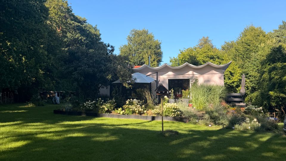 Großer Garten mit weitläufiger Rasenfläche, Blumen- und Strauchbeeten sowie hohen Bäumen am Rand. Im Hintergrund steht ein flaches, modernes Gebäude mit markant gewelltem Dach und einer Terrasse mit Sonnenschirmen.