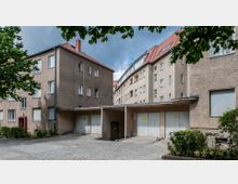 Wohnhof mit mehrstöckigen Wohnhäusern in einfacher Putzfassade und rotem Ziegeldach. Im Vordergrund ein kleiner Innenhof mit Garagen und Pflastersteinen, umrandet von Büschen und Bäumen.