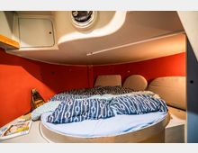 Kleiner Schlafbereich in einer Bootskabine mit einem runden Bett, roten Wänden und blauer Bettwäsche mit weißen Mustern. Oberhalb befindet sich ein kleines Bullauge, daneben sind Ablageflächen sowie persönliche Gegenstände sichtbar.