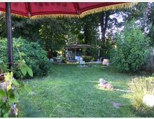 Garten mit Rasenfläche, umgeben von dichtem, grünem Gebüsch und hohen Bäumen. Im Hintergrund ist eine kleine Hütte mit Veranda, davor stehen bunte Gartenstühle; im Vordergrund liegt ein Hund unter einem roten Sonnenschirm.