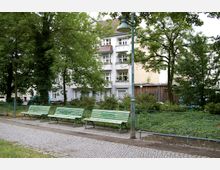 Städtischer Parkbereich mit gepflastertem Weg, drei grünen Bänken und hohen Bäumen im Vordergrund. Im Hintergrund befindet sich ein mehrstöckiges Wohnhaus mit weißen Fassaden und Balkonen sowie ein Zaun und eine Grünanlage.
