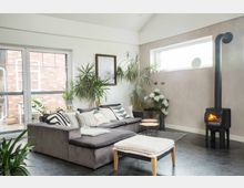 Modernes Wohnzimmer mit einer grauen Eckcouch, dekorativen Kissen und einem kleinen Hocker. Im Raum befinden sich zahlreiche Zimmerpflanzen, ein an der Wand montierter Holzbrennofen und große Fenster, die viel Tageslicht einlassen.