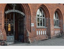 Außenansicht eines historischen Backsteingebäudes mit Spitzbögen und großen Fenstern, darunter der Eingang, der durch ein schmiedeeisernes Tor mit der Aufschrift „Hallesches Haus“ markiert ist. Daneben steht ein bemalter Wandbereich, und vor dem Gebäude sind Fahrräder abgestellt.