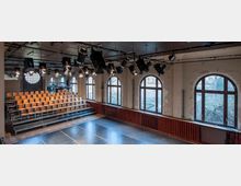 Kleiner Veranstaltungssaal mit Holzbühne, gestuften Sitzreihen aus Holzstühlen und großen Rundbogenfenstern. Die Wände sind leicht abgeblättert, und an der Decke befinden sich mehrere Scheinwerfer.