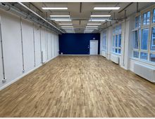 Ein leerer, rechteckiger Raum mit hellbraunem Parkettboden, weißen Wänden und einer dunkelblauen Akzentwand im Hintergrund. Oben an der Decke verlaufen sichtbare Lüftungsrohre und Hängelampen, während die rechte Wand von großen Fensterfronten mit Heizkörpern darunter dominiert wird.