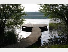 Holzsteg, der geschwungen über das Wasser eines ruhigen Sees führt, umgeben von dichtem Baumbewuchs und Schilf am Ufer. Im Hintergrund sind bewaldete Hügel zu sehen, die sich im Wasser spiegeln.