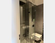 Modernes Badezimmer mit grauen Fliesen und einer bodengleichen Glasdusche mit schwarzem Rahmen und Regendusche. Rechts ist ein wandhängendes WC zu sehen sowie eine eingelassene Wandnische für Pflegeprodukte.