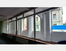 Innenansicht eines Gebäudes mit großen Glasfenstern und Blick auf die Fassade eines gegenüberliegenden Hauses im Gründerzeitstil. Vor den Fenstern stehen weiße Heizkörper, und an einer Scheibe klebt ein blauer Hinweis auf eine räumliche Veränderung.