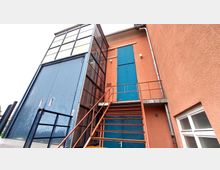 Außenansicht eines Gebäudes mit orangefarbener Fassade und blauen Türen. Eine Metalltreppe mit roten Geländern führt zu einem erhöhten Eingang, daneben ein hoher Bereich mit großen Glasfenstern und einer blauen Schiebetür.