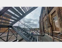 Metall-Treppenaufgang an der Außenseite eines Backsteingebäudes, mit Blick auf einen Hinterhof mit geparkten Fahrzeugen und einem Mobilfunkmast in der Ferne unter bewölktem Himmel.