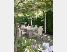 Garten mit einer überdachten Sitzecke, bestehend aus einem hellen Tisch und mehreren Stühlen auf einem gepflasterten Untergrund. Die Überdachung ist mit Pflanzen und Blättern bewachsen, im Hintergrund ist eine dichte Hecke zu sehen.