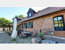 Ziegelsteingebäude mit schwarzen Fensterläden und einer überdachten Terrasse, auf der Holzbänke und Korbstühle um Tische auf Kopfsteinpflaster stehen. Im Vordergrund ein Sonnenschirm und Blumendekoration, im Hintergrund grüne Bäume.