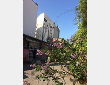 Innenhof zwischen mehrstöckigen Gebäuden mit großen Wandgemälden auf den Fassaden und einer verglasten Werkstattfront. Im Vordergrund stehen blühende Sträucher und weitere Begrünung; über dem Hof hängen bunte Wimpelketten vor blauem Himmel.