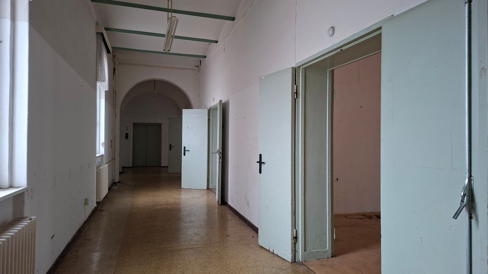 Langer, leerer Flur in einem älteren Gebäude mit hohen weißen Wänden, Fensterreihe links und mehreren offenen, hellblauen Türen zu Nebenräumen. Deckenleuchten hängen an grünen Trägern, am Ende des Ganges befindet sich ein Rundbogen mit einer weiteren Tür.